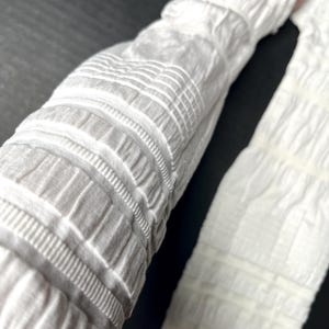 May include: A pair of white arm sleeves featuring a textured, ruched fabric design with alternating horizontal bands of gathered material and smooth, flat sections. The sleeves are constructed from a lightweight, stretchy, and opaque white textile.