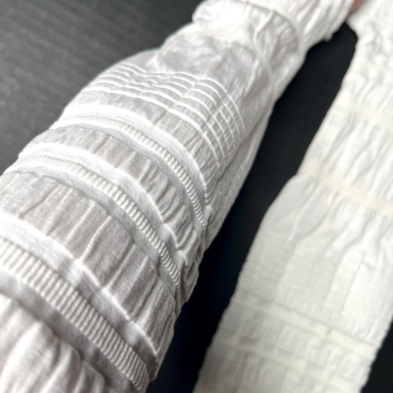May include: A pair of white arm sleeves featuring a textured, ruched fabric design with alternating horizontal bands of gathered material and smooth, flat sections. The sleeves are constructed from a lightweight, stretchy, and opaque white textile.