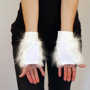 May include: A pair of white fingerless gloves featuring a smooth fabric center panel and long, fluffy white faux fur trim along the sides. The gloves have a simple, clean design with a solid white elasticated cuff at the wrist for a secure fit.