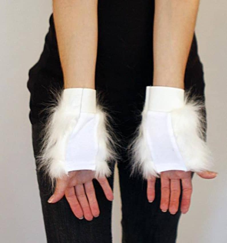 May include: A pair of white fingerless gloves featuring a smooth fabric center panel and long, fluffy white faux fur trim along the sides. The gloves have a simple, clean design with a solid white elasticated cuff at the wrist for a secure fit.