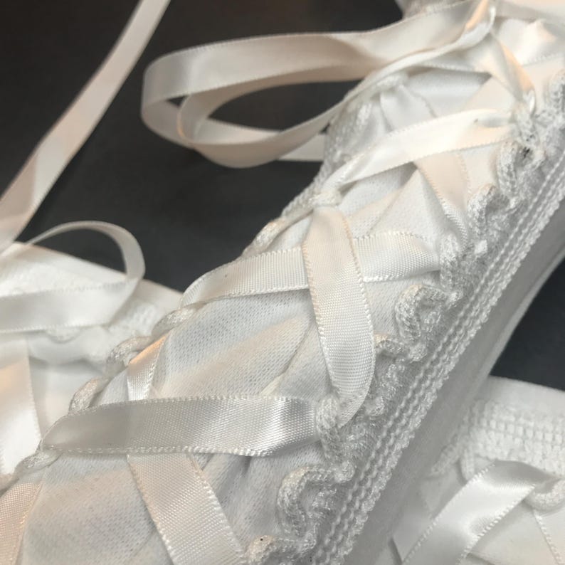 May include: A white fabric item featuring a crisscross lacing pattern created with smooth satin ribbon. The edges are finished with a delicate, scalloped crochet trim. The white material has a fine, textured weave, and long ribbon ends extend outward.