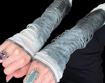 May include: A pair of long, layered arm warmers in shades of gray and white. The fabric has a distressed, shredded appearance, with a delicate, lacy texture. The cuffs are trimmed with black stitching. The arm warmers are shown against a black background.