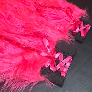 May include: Bright pink faux fur with matching satin ribbon accents. Two black wrist cuffs with pink ribbon lacing are visible. The image is set against a dark background.