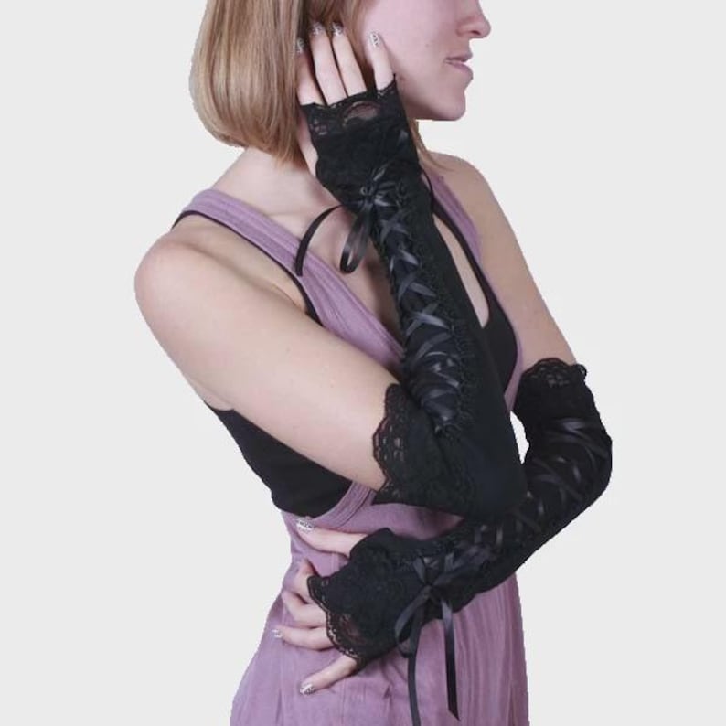 May include: A pair of black fingerless gloves featuring intricate lace trim at the edges and a corset-style lace-up design with thin black ribbons running down the length of the forearm. The gloves extend past the elbow and include small bows.