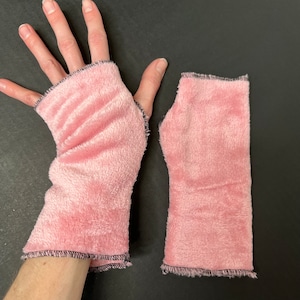 Puede incluir: Un par de calientamanos de felpa rosa con costuras negras. Los calientamanos se muestran colocados sobre una superficie oscura.