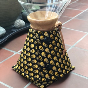 Op de afbeelding: Een zwarte en gouden stoffen koffiefilterhouder met een schedelpatroon. De houder heeft een houten ring en een glazen karaf.