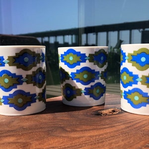 Puede incluir: Tres tazas de cerámica blanca con un patrón geométrico en tonos de azul, verde y blanco. Las tazas tienen un asa en el lado derecho.