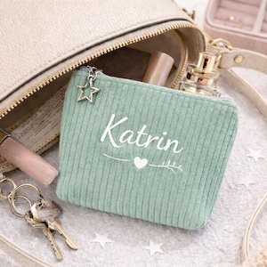 Könnte beinhalten: Ein hellgrünes Cord-Etui mit dem Namen "Katrin" in weißer Schrift, einem Sternanhänger und einem Herzdesign. Das Etui befindet sich in einer beigen Handtasche mit goldenem Reißverschluss, neben Schlüsseln, Make-up und Parfüm.