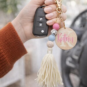 May include: A personalised keychain with a wooden round tag that says "Katrin" in pink script. The keychain has a gold clasp, colourful beads, and a cream-coloured tassel. A car key fob is also attached.