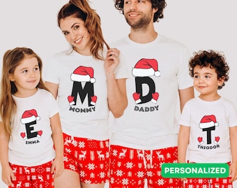 May include: Matching family Christmas pajamas. White t-shirts with a Santa hat and initial design, paired with red shorts featuring a white snowflake and reindeer pattern. The word "PERSONALIZED" is visible.