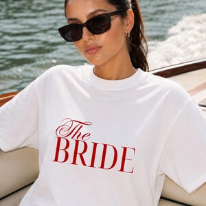 Op de afbeelding: Een wit T-shirt met korte mouwen en een ronde hals, voorzien van de tekst "The BRIDE" in een opvallend rood schreeflettertype op de borst. Het shirt heeft een relaxte pasvorm met een standaard zoom en is gemaakt van gladde, witte stof.