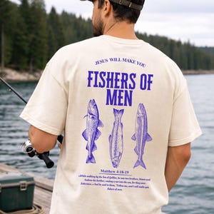 May include: Cream-colored t-shirt with the text "JESUS WILL MAKE YOU FISHERS OF MEN" in blue. Below the text are three blue fish illustrations. The shirt also features a Bible verse. The person in the image is holding a fishing rod.