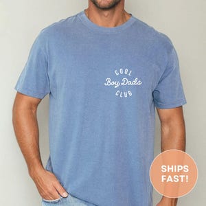 May include: A blue short-sleeved t-shirt with the text "COOL Boy Dads CLUB" in white script on the upper left chest. The shirt is a solid blue color and has a crew neck.