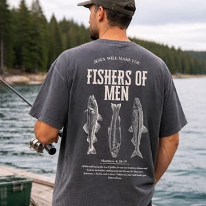May include: Dark gray t-shirt with the text "JESUS WILL MAKE YOU FISHERS OF MEN" and three fish illustrations. The shirt also includes the Bible verse Matthew 4:18-19. The person is holding a fishing rod.