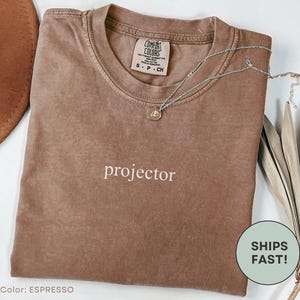 May include: Espresso-colored t-shirt with the word "projector" in white script. A silver necklace with a pendant rests on the shirt. The shirt is folded and has a tag that says "Comfort Colors".