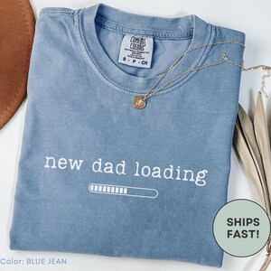 May include: Blue jean Comfort Colors t-shirt with the text "new dad loading" in white. A gold necklace with a pendant rests on the shirt. The shirt has a crew neck and short sleeves.