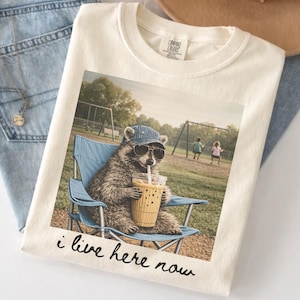 K&ouml;nnte beinhalten: Cremefarbenes Comfort Colors T-Shirt mit einem Aufdruck eines Waschb&auml;ren mit Sonnenbrille und Jeansm&uuml;tze, der in einem blauen Klappstuhl sitzt und aus einem Becher trinkt. Der Text "i live here now" ist darunter gedruckt. Eine Schaukel ist im Hintergrund.