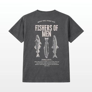 Puede incluir: Camiseta gris oscuro con el texto "JESUS WILL MAKE YOU FISHERS OF MEN" y tres ilustraciones de peces. El texto "Matthew 4:18-19" est&aacute; debajo. La camiseta tiene un aspecto vintage y desgastado.