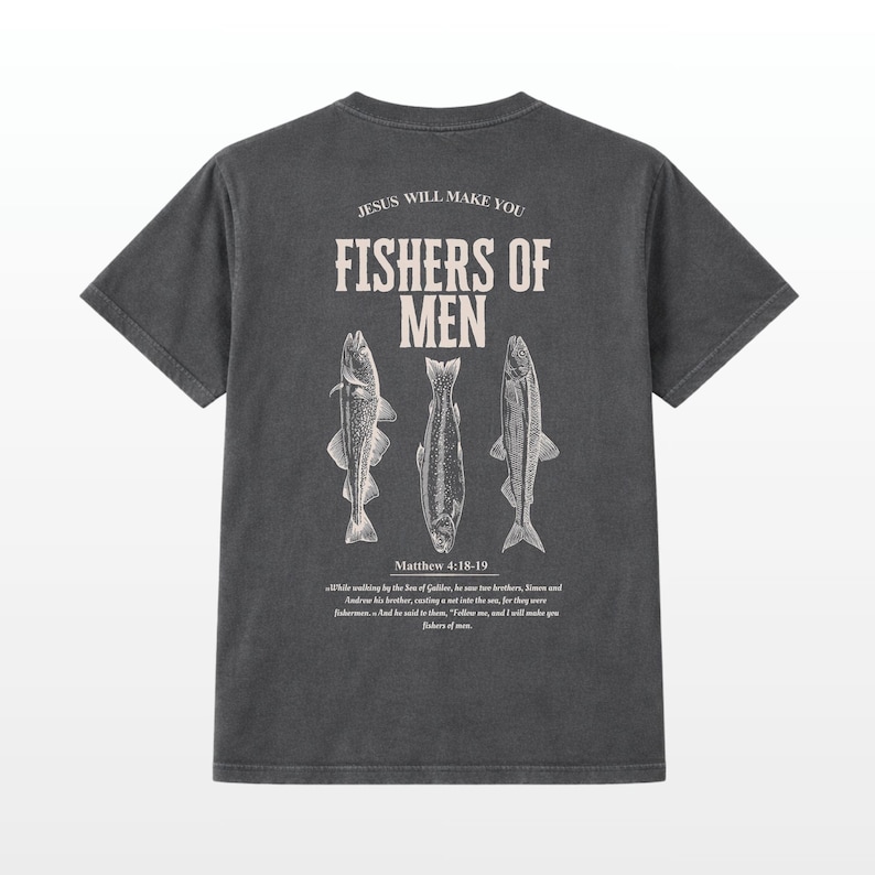 Puede incluir: Camiseta gris oscuro con el texto "JESUS WILL MAKE YOU FISHERS OF MEN" y tres ilustraciones de peces. El texto "Matthew 4:18-19" est&aacute; debajo. La camiseta tiene un aspecto vintage y desgastado.