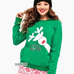 May include: A green sweatshirt with a white reindeer graphic and the text "#TEAM RUDOLPH". The person is wearing a black beanie and red and white patterned pants. The sweatshirt is a festive holiday design.