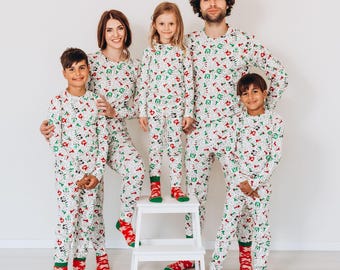 Könnte beinhalten: Eine fünfköpfige Familie trägt passende weiße Pyjamas mit einem festlichen Weihnachtsdruck. Die Pyjamas haben ein rot-grün-weißes Muster mit verschiedenen Weihnachtssymbolen. Die Familie steht vor einer weißen Wand und die Kinder tragen passende rot-grüne Socken.