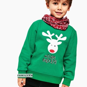 May include: A green sweatshirt featuring a white reindeer graphic with a red nose and the text "#TEAM RUDOLPH". The child is wearing a red and white patterned neck warmer. The sweatshirt is a festive holiday garment.