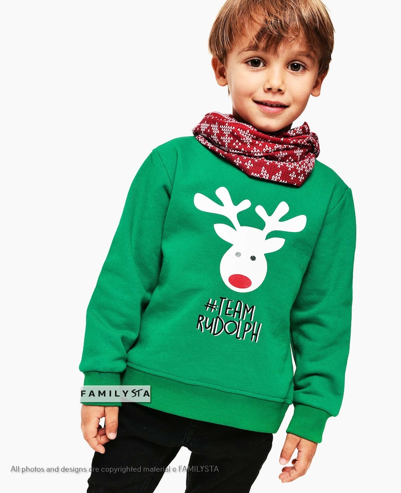 May include: A green sweatshirt featuring a white reindeer graphic with a red nose and the text "#TEAM RUDOLPH". The child is wearing a red and white patterned neck warmer. The sweatshirt is a festive holiday garment.