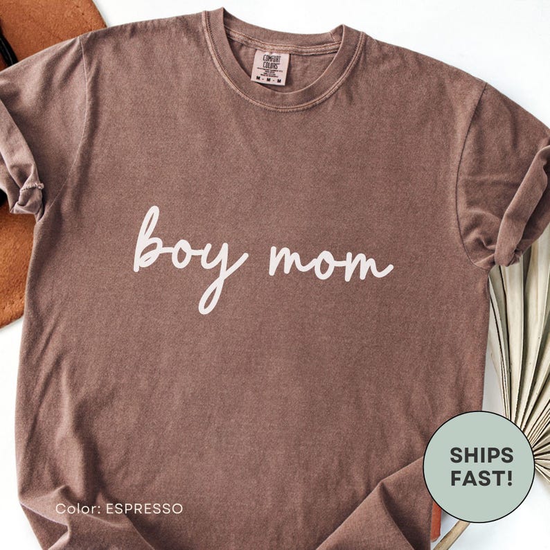 K&ouml;nnte beinhalten: Espressofarbenes T-Shirt mit dem wei&szlig;en Schriftzug "boy mom". Das Shirt hat einen Rundhalsausschnitt und kurze &Auml;rmel. Die Farbe ist unten links mit "espresso" gekennzeichnet. Ein runder Aufkleber unten rechts zeigt "SHIPS FAST!"