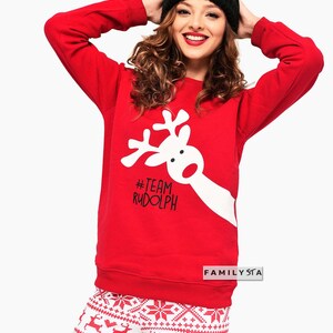 May include: Red sweatshirt with a white reindeer graphic and the text "#TEAM RUDOLPH". The model is wearing a black beanie and white and red patterned pants. A festive outfit for the holiday season.
