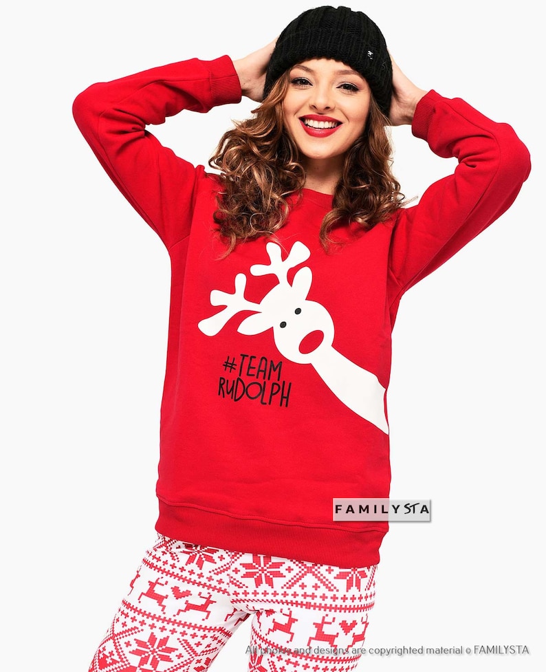 May include: Red sweatshirt with a white reindeer graphic and the text "#TEAM RUDOLPH". The model is wearing a black beanie and white and red patterned pants. A festive outfit for the holiday season.