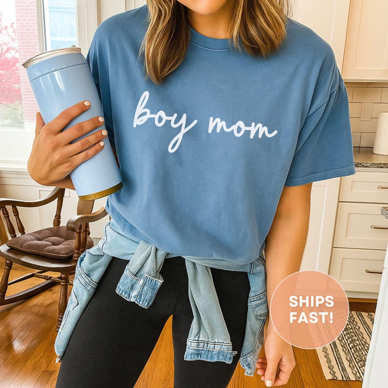 K&ouml;nnte beinhalten: Hellblaues T-Shirt mit dem wei&szlig;en Schriftzug "boy mom". Das Shirt wird mit schwarzen Leggings und einer um die Taille gebundenen Jeansjacke getragen. Ein hellblauer Becher mit goldenem Boden wird in der Hand gehalten.