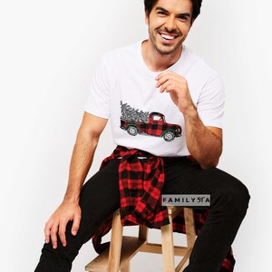 May include: White t-shirt with a red and black plaid pickup truck carrying a Christmas tree. The person is wearing black jeans and a red and black plaid shirt tied around their waist. The text "FAMILYSTA" is visible on the shirt.