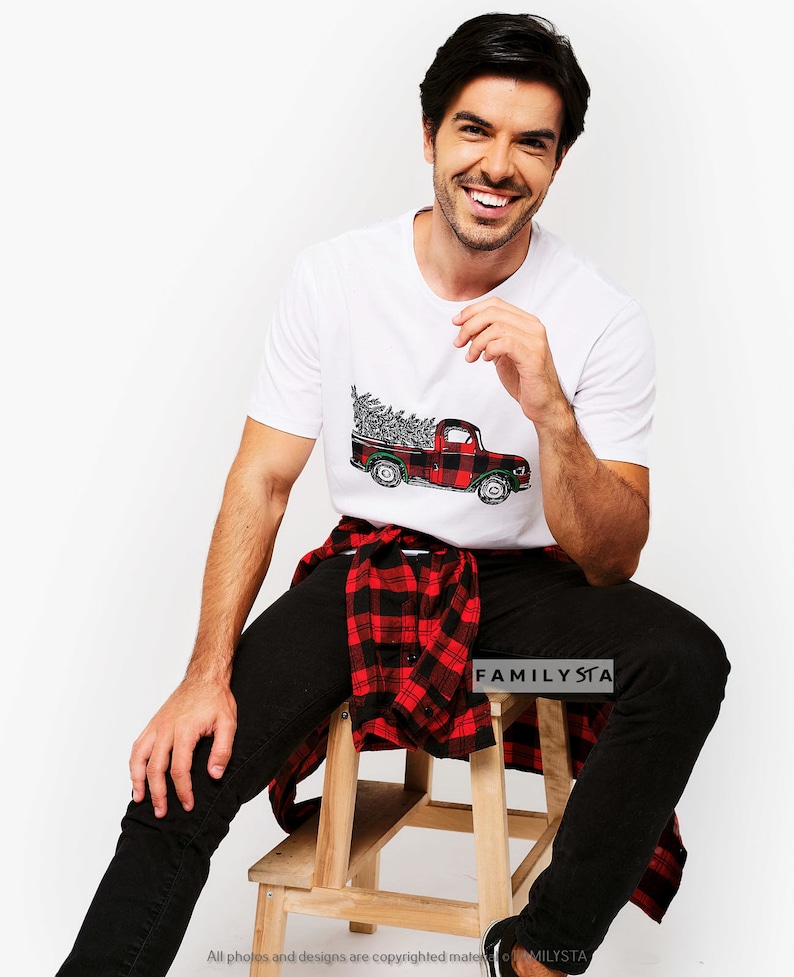 May include: White t-shirt with a red and black plaid pickup truck carrying a Christmas tree. The person is wearing black jeans and a red and black plaid shirt tied around their waist. The text "FAMILYSTA" is visible on the shirt.