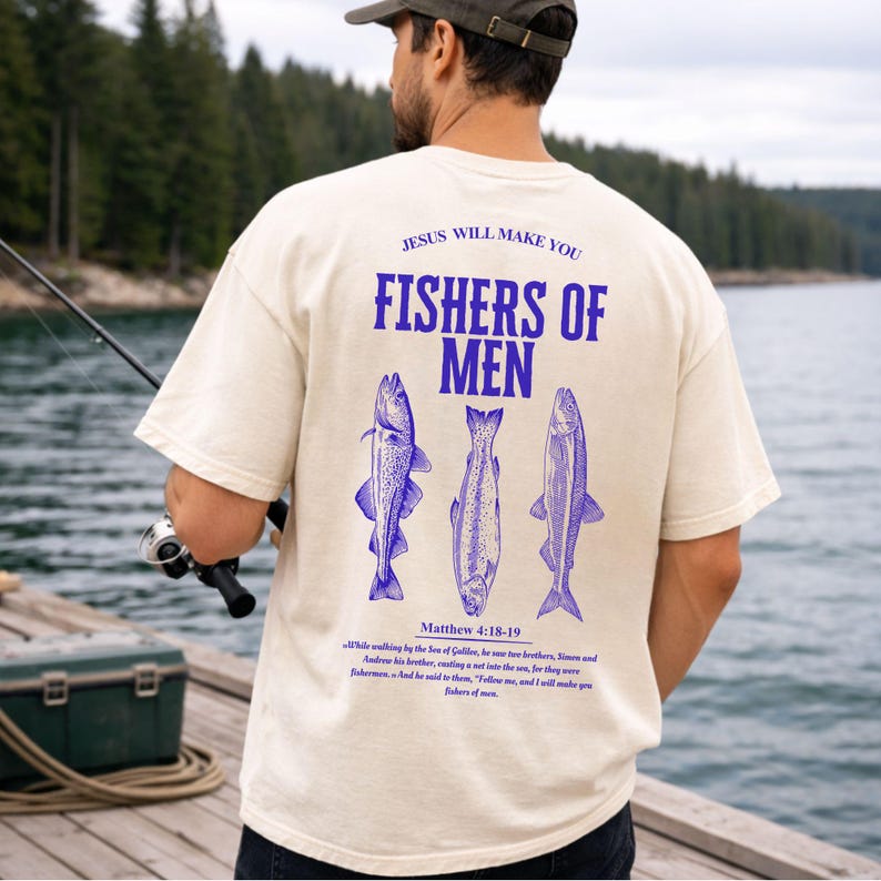 May include: Cream-colored t-shirt with the text "JESUS WILL MAKE YOU FISHERS OF MEN" in blue. Below the text are three blue fish illustrations. The shirt also features a Bible verse. The person in the image is holding a fishing rod.