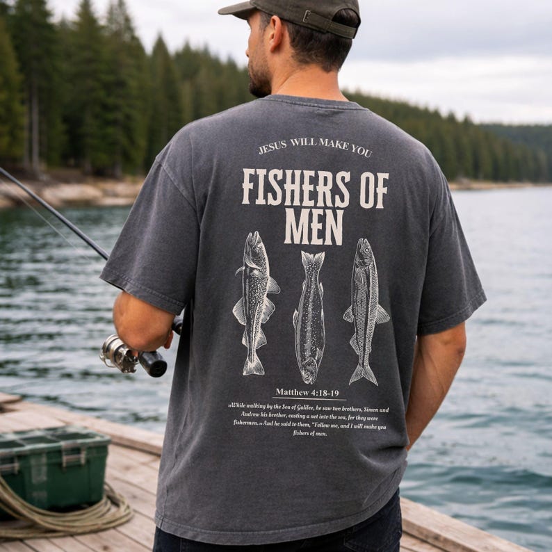 May include: Dark gray t-shirt with the text "JESUS WILL MAKE YOU FISHERS OF MEN" and three fish illustrations. The shirt also includes the Bible verse Matthew 4:18-19. The person is holding a fishing rod.