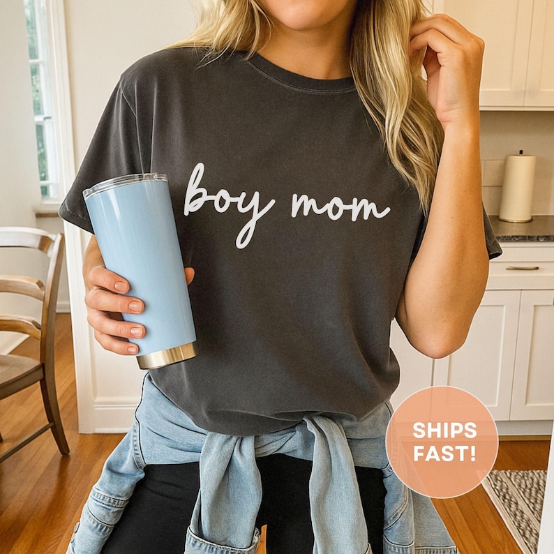 K&ouml;nnte beinhalten: Dunkelgraues T-Shirt mit dem Schriftzug "boy mom" in wei&szlig;er Schrift. Ein hellblauer Becher mit goldenem Boden wird in der Hand gehalten. Das Shirt wird mit einer schwarzen Hose und einer um die Taille gebundenen Jeansjacke kombiniert.