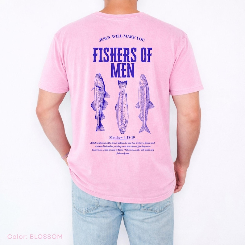 Puede incluir: Camiseta rosa claro con el texto azul "JESUS WILL MAKE YOU FISHERS OF MEN" y tres ilustraciones de peces azules. El texto "Matthew 4:18-19" tambi&eacute;n est&aacute; impreso. La camiseta es de color pastel claro.