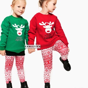 May include: Two young children wearing matching Christmas outfits. One child is wearing a green sweatshirt and the other a red sweatshirt, both with a white reindeer design and the text "#TEAM RUDOLPH". They are wearing red and white patterned leggings and black boots.