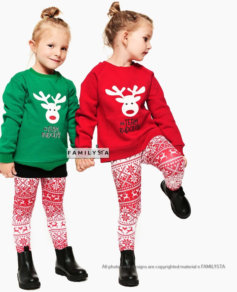 May include: Two young children wearing matching Christmas outfits. One child is wearing a green sweatshirt and the other a red sweatshirt, both with a white reindeer design and the text "#TEAM RUDOLPH". They are wearing red and white patterned leggings and black boots.