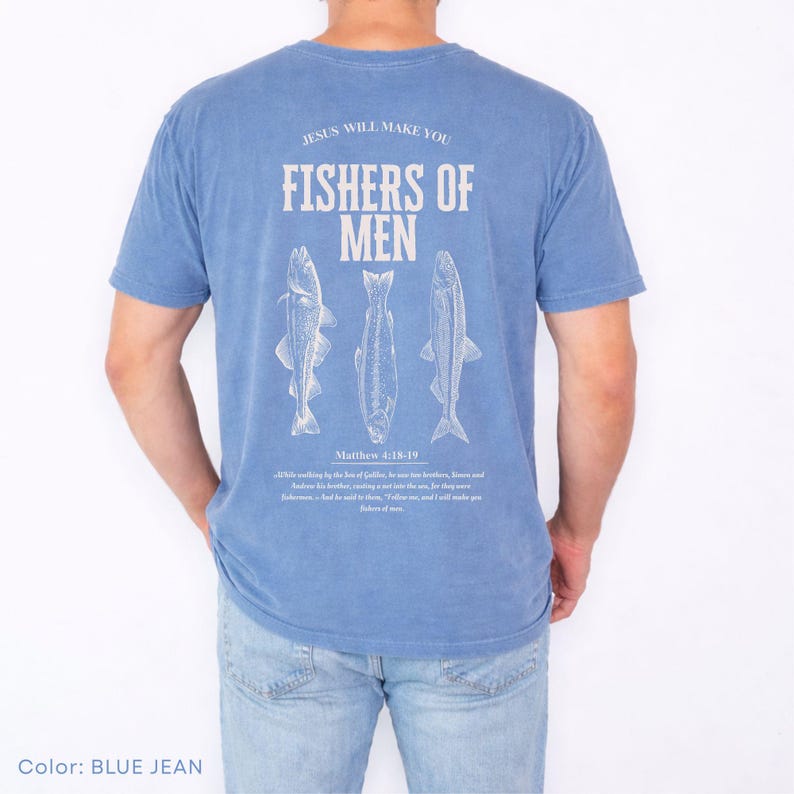 Puede incluir: Camiseta azul vaquero de manga corta con el texto "JESUS WILL MAKE YOU FISHERS OF MEN" y tres ilustraciones de peces. El texto "Matthew 4:18-19" est&aacute; impreso debajo.