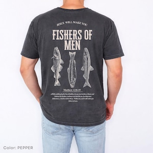 Puede incluir: Camiseta gris oscuro con el texto "JESUS WILL MAKE YOU FISHERS OF MEN" y tres ilustraciones de peces. El texto "Matthew 4:18-19" y un vers&iacute;culo b&iacute;blico est&aacute;n impresos debajo. Una camiseta informal de manga corta.