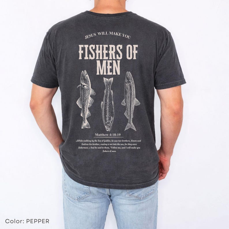 Puede incluir: Camiseta gris oscuro con el texto "JESUS WILL MAKE YOU FISHERS OF MEN" y tres ilustraciones de peces. El texto "Matthew 4:18-19" y un vers&iacute;culo b&iacute;blico est&aacute;n impresos debajo. Una camiseta informal de manga corta.