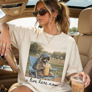 K&ouml;nnte beinhalten: Cremefarbenes T-Shirt mit einem Aufdruck eines Waschb&auml;ren mit Sonnenbrille und Hut, der in einem blauen Stuhl sitzt und ein Getr&auml;nk h&auml;lt. Der Aufdruck enth&auml;lt den Text "i live here now". Das Shirt wird von einer Person im Auto getragen.