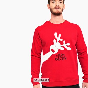 May include: A red sweatshirt with a white reindeer graphic and the text "#TEAM RUDOLPH". The sweatshirt is a crew neck style and is made of a soft material. The image also includes the text "FAMILYSTA".