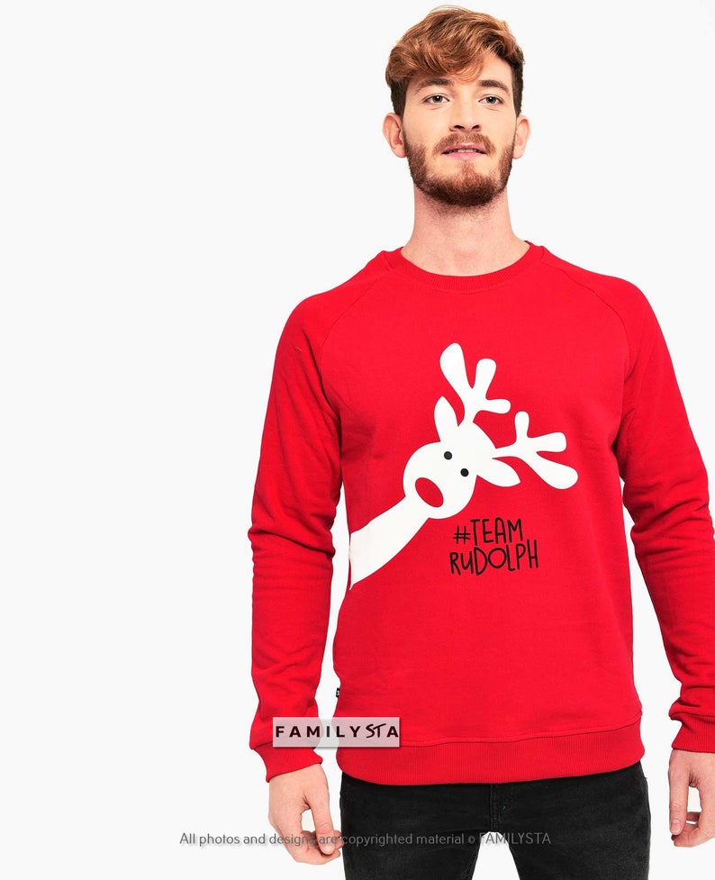 May include: A red sweatshirt with a white reindeer graphic and the text "#TEAM RUDOLPH". The sweatshirt is a crew neck style and is made of a soft material. The image also includes the text "FAMILYSTA".