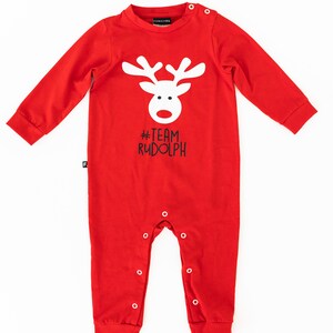 May include: A red baby onesie with long sleeves and snap closures. The onesie features a white reindeer face graphic with the text "#TEAM RUDOLPH" in black. The garment is made of a soft material.