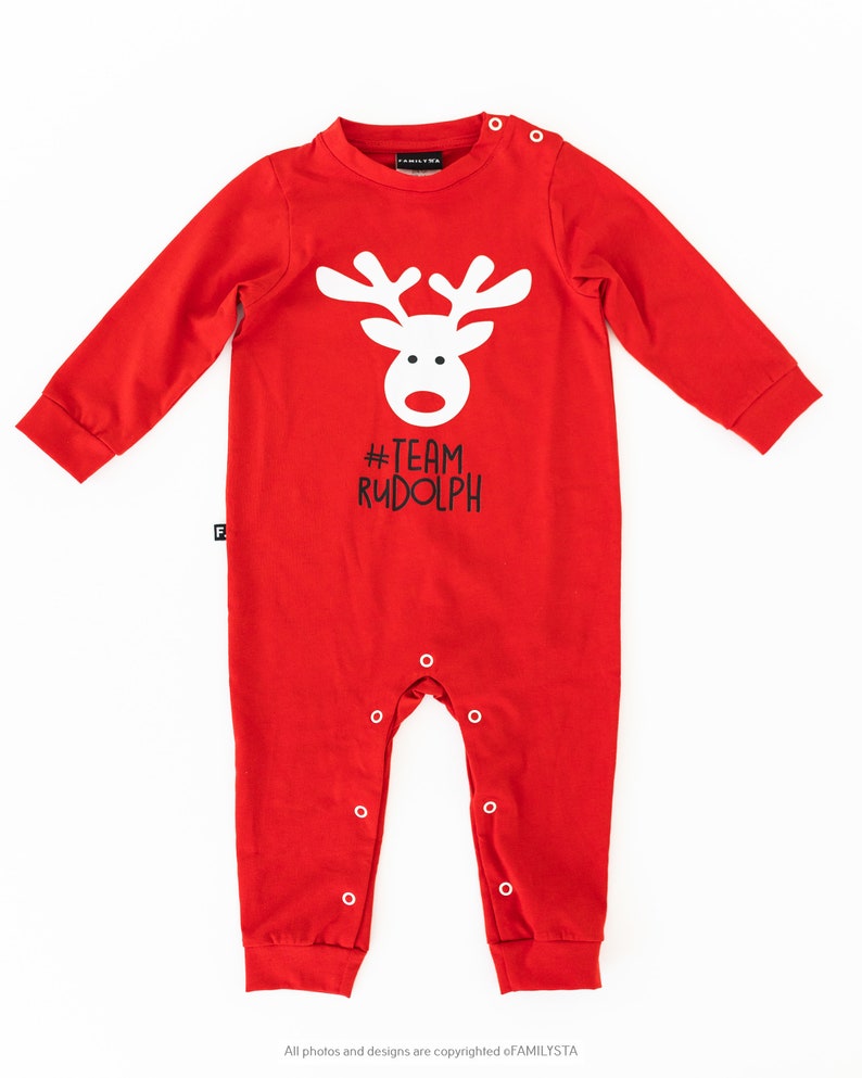 May include: A red baby onesie with long sleeves and snap closures. The onesie features a white reindeer face graphic with the text "#TEAM RUDOLPH" in black. The garment is made of a soft material.