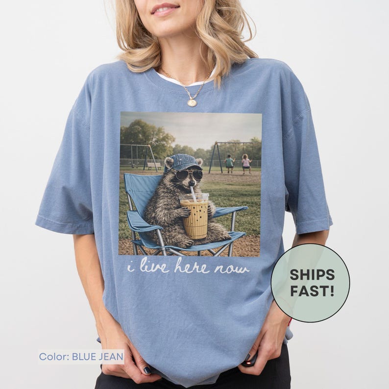 K&ouml;nnte beinhalten: Blaues Jeans-T-Shirt mit einer Grafik eines Waschb&auml;ren mit Sonnenbrille und Hut, der in einem blauen Stuhl sitzt und ein Getr&auml;nk h&auml;lt. Der Text "i live here now" steht darunter.