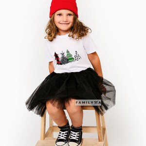 May include: A young person wearing a white t-shirt with a Christmas tree design, a black tulle skirt, red beanie, and black sneakers. The t-shirt has the text "FAMILYSTA" on the bottom.