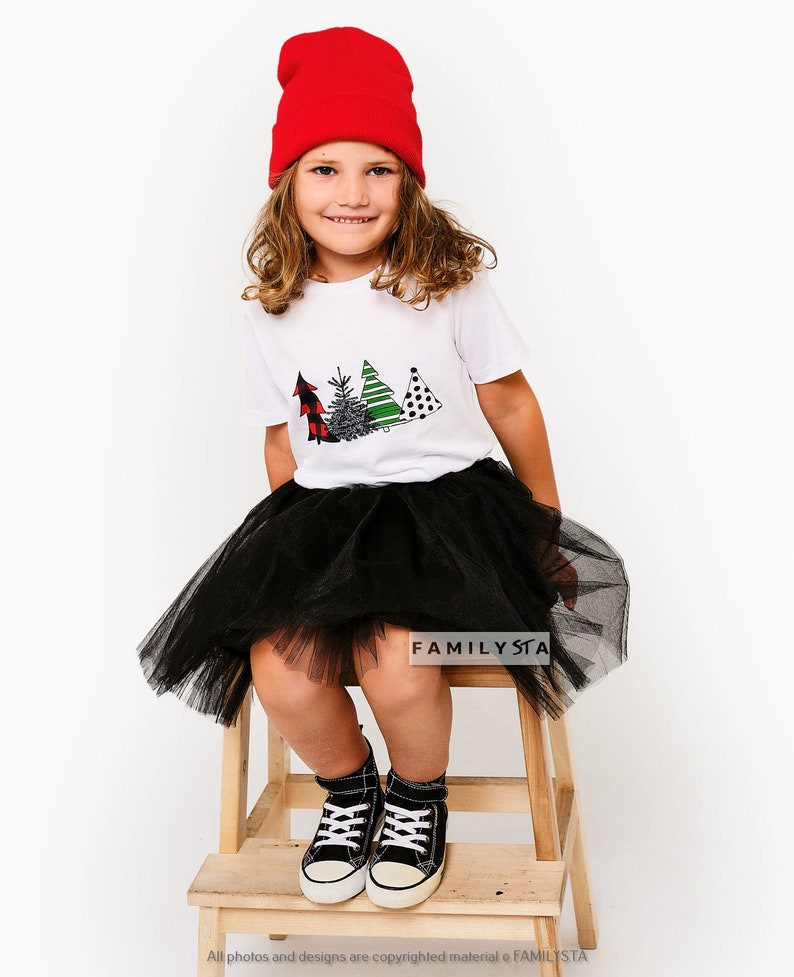 May include: A young person wearing a white t-shirt with a Christmas tree design, a black tulle skirt, red beanie, and black sneakers. The t-shirt has the text "FAMILYSTA" on the bottom.